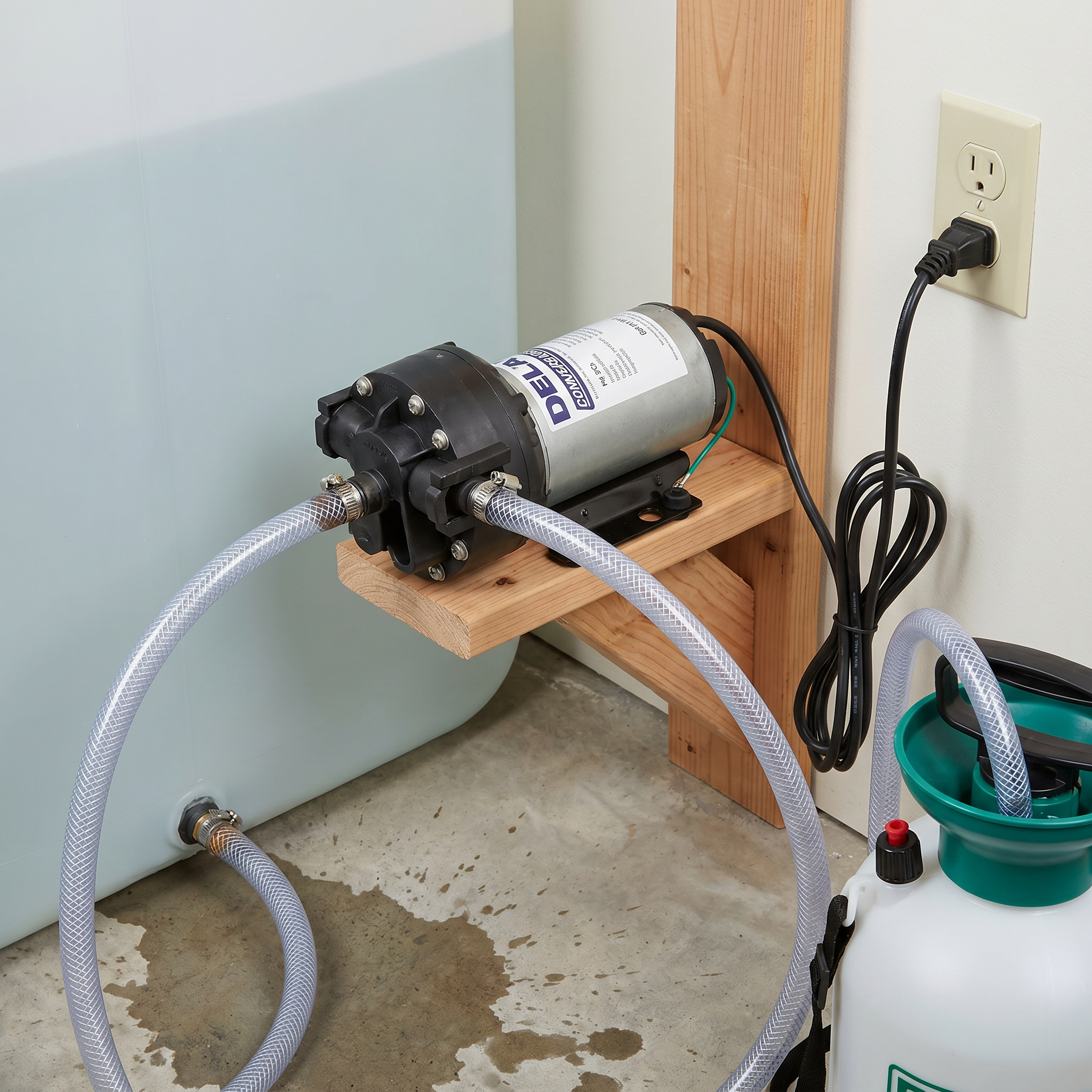The electric diaphragm transfer pump mounted on a wooden wall bracket, plugged into a wall outlet and connected via clear braided hoses to pull fluid from a white chemical sprayer tank.