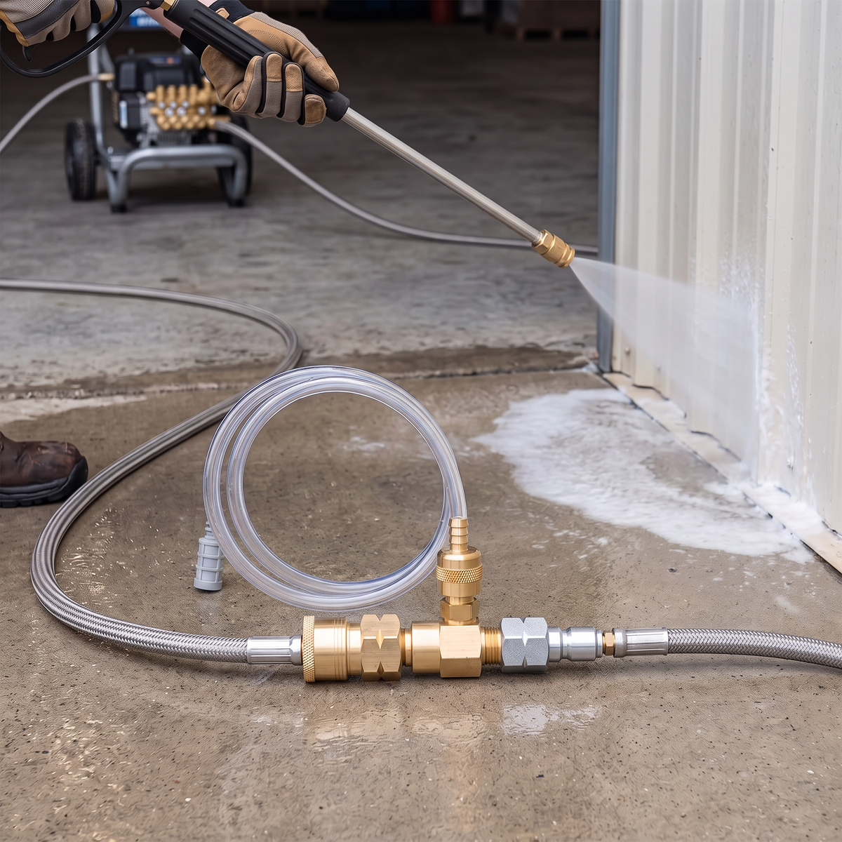 Brass pressure washer downstream chemical injector connected inline to a high-pressure hose with a coiled clear suction tube, showing an operator spraying soap solution against a metal wall in the background.